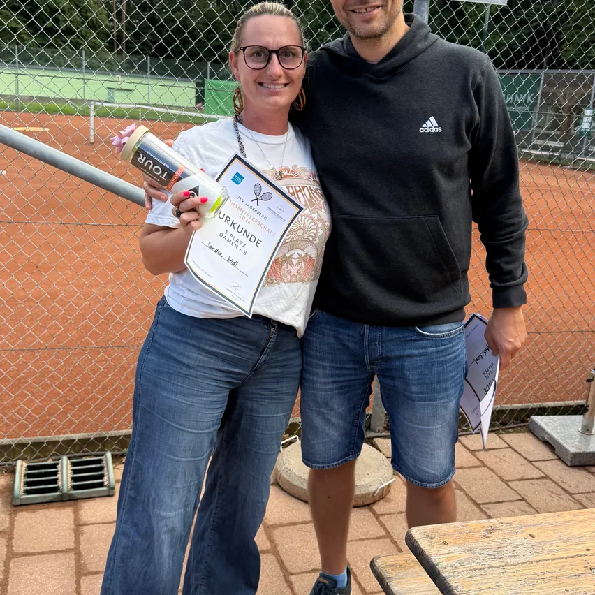 Ein Mann und eine Frau stehen lächelnd zusammen draußen, mit einem Tennisplatz im Hintergrund. Die Frau hält ein aufgerolltes Zertifikat und eine Trophäe.