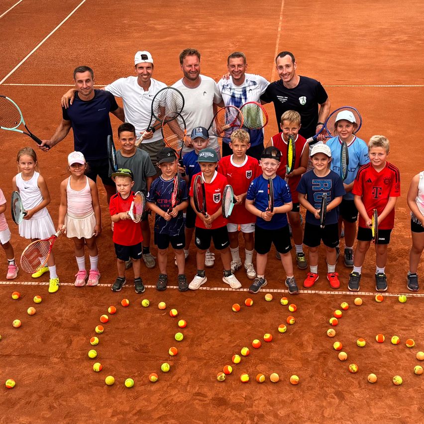 A group of people pose for a photo on a tennis court with tennis balls arranged in the shape of a heart. Some are holding rackets and wearing hats and sneakers.
