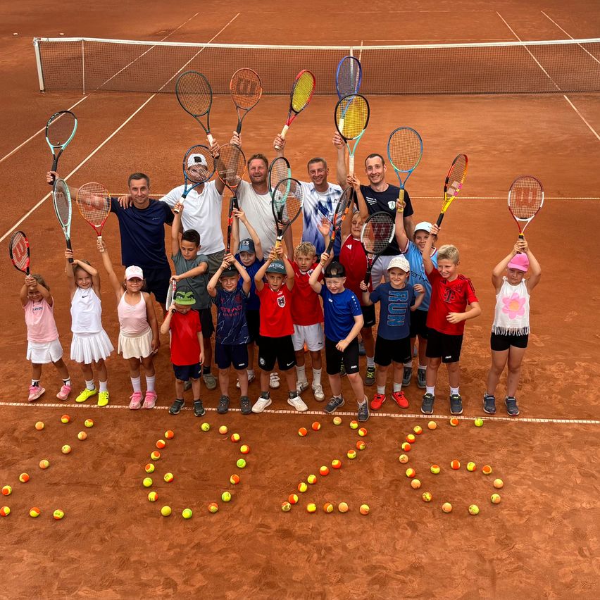 A group of young children and adults are posing for a photo on a tennis court, holding tennis rackets. The court is surrounded by tennis balls arranged in a pattern.