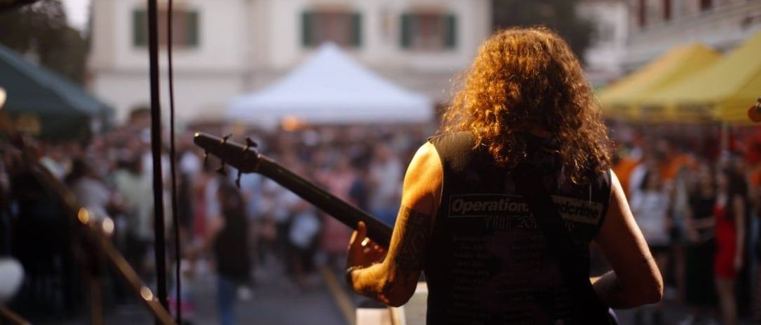 Ein Gitarrist spielt auf der Bühne vor einer großen Menge. Ein Zelt und Gebäude sind sichtbar.