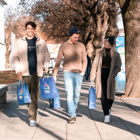 Drei Personen gehen mit blauen Einkaufstüten auf einem Bürgersteig. Der Mann auf der linken Seite trägt eine beige Jacke und Jeans. Der Mann in der Mitte trägt ein braunes Hemd und Jeans. Die Frau auf der rechten Seite trägt einen beigefarbenen Mantel und schwarze Leggings.
