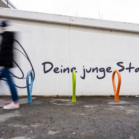 Eine Person geht an einer weißen Wand mit farbenfrohen Graffiti vorbei. Die Graffiti lesen 'Deine junge Stadt'. Es gibt drei farbenfrohe Fahrradständer auf dem Boden.
