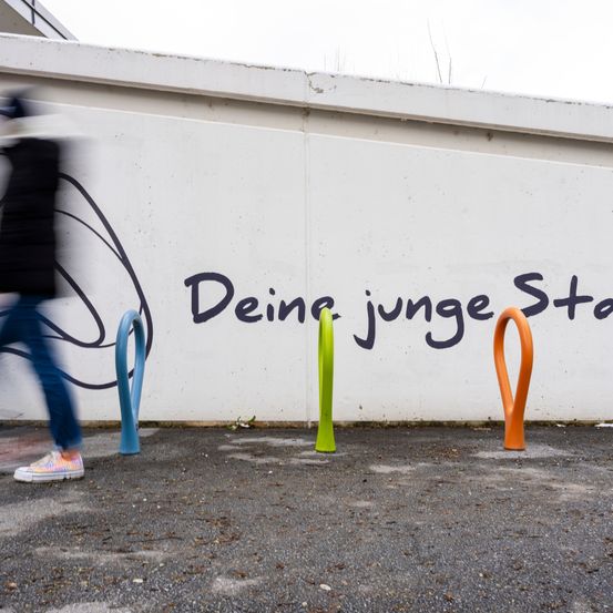 Eine Person geht an einer weißen Wand mit farbenfrohen Graffiti vorbei. Die Graffiti lesen 'Deine junge Stadt'. Es gibt drei farbenfrohe Fahrradständer auf dem Boden.