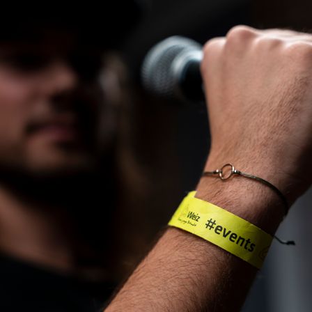Ein Mann mit einem gelben Armband, das mit 'Weiz #events' beschriftet ist, hält ein Mikrofon, sein Gesicht ist teilweise im Hintergrund zu sehen.