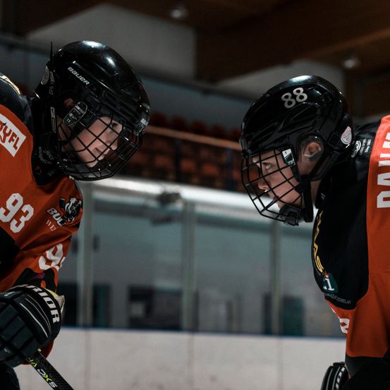 Zwei Eishockeyspieler stehen sich in einer Eishalle gegenüber. Beide tragen schwarze Helme und orangefarbene Trikots mit Nummern. Einer hat '33' und der andere '88'.