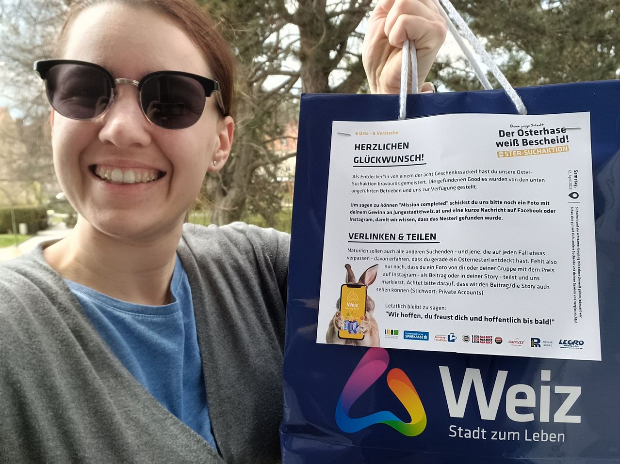 Eine Frau mit Sonnenbrille hält einen blauen Beutel, auf dem 'Herzlichen Gluckwunsch!' und 'Weihnachtswunsch!' steht. Der Beutel hat auch Logos und Text, der 'Wir hoffen, du freust dich und hoffentlich bis bald' lautet.