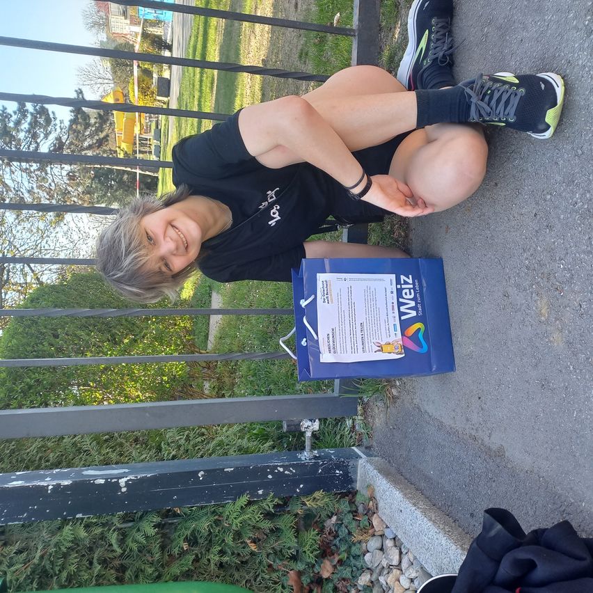 Eine lächelnde Frau posiert mit einer blauen Box mit der Aufschrift 'Weiz' neben ihr, während sie auf einem Zaun mit einem Metallgeländer in einem Park sitzt.