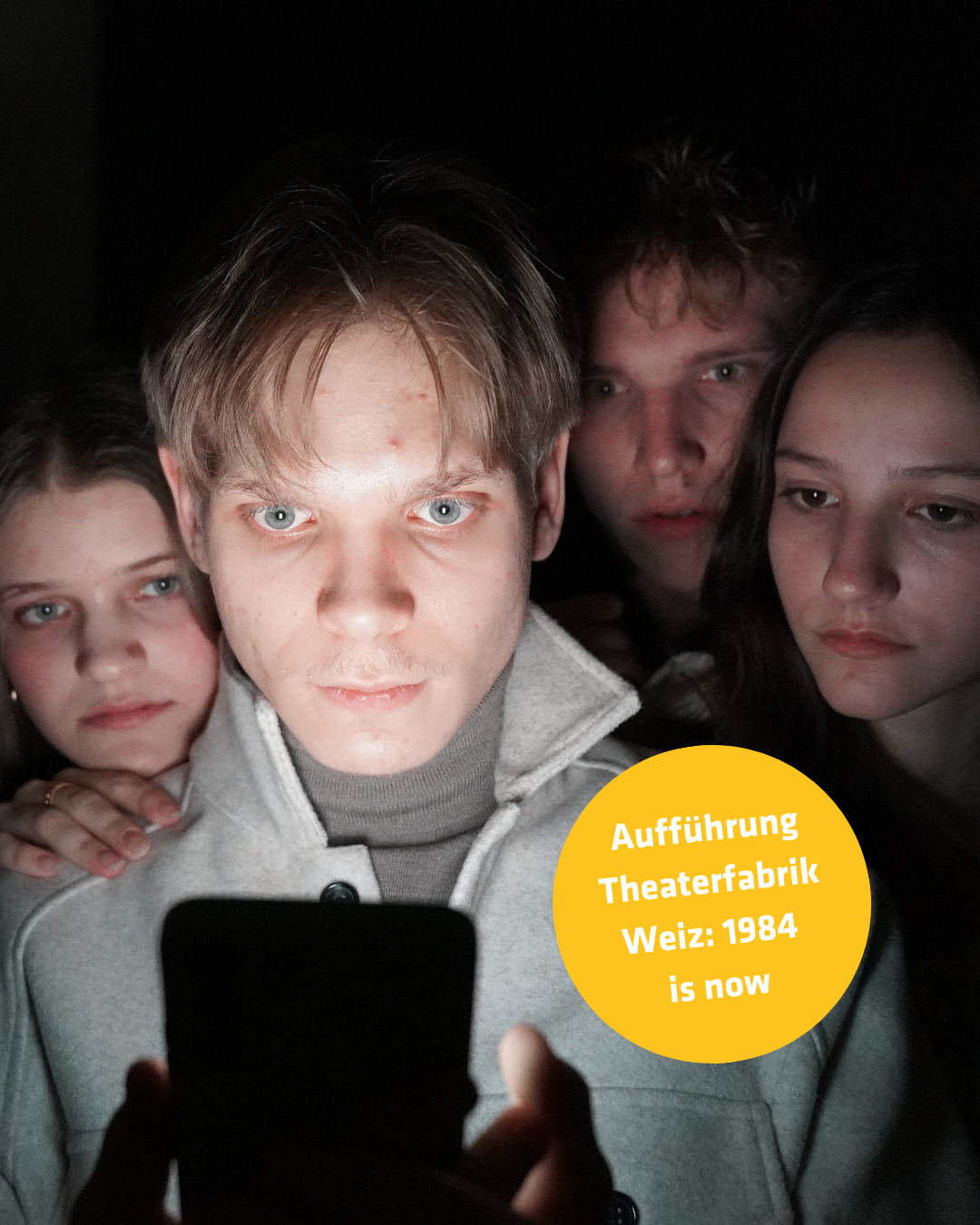 Vier junge Menschen sind in einem schwach beleuchteten Raum. Der Mann in der Mitte hält ein Handy und schaut darauf. Der Text auf dem Bild lautet: 'Aufführung Theaterfabrik Weiz: 1984 ist jetzt.'