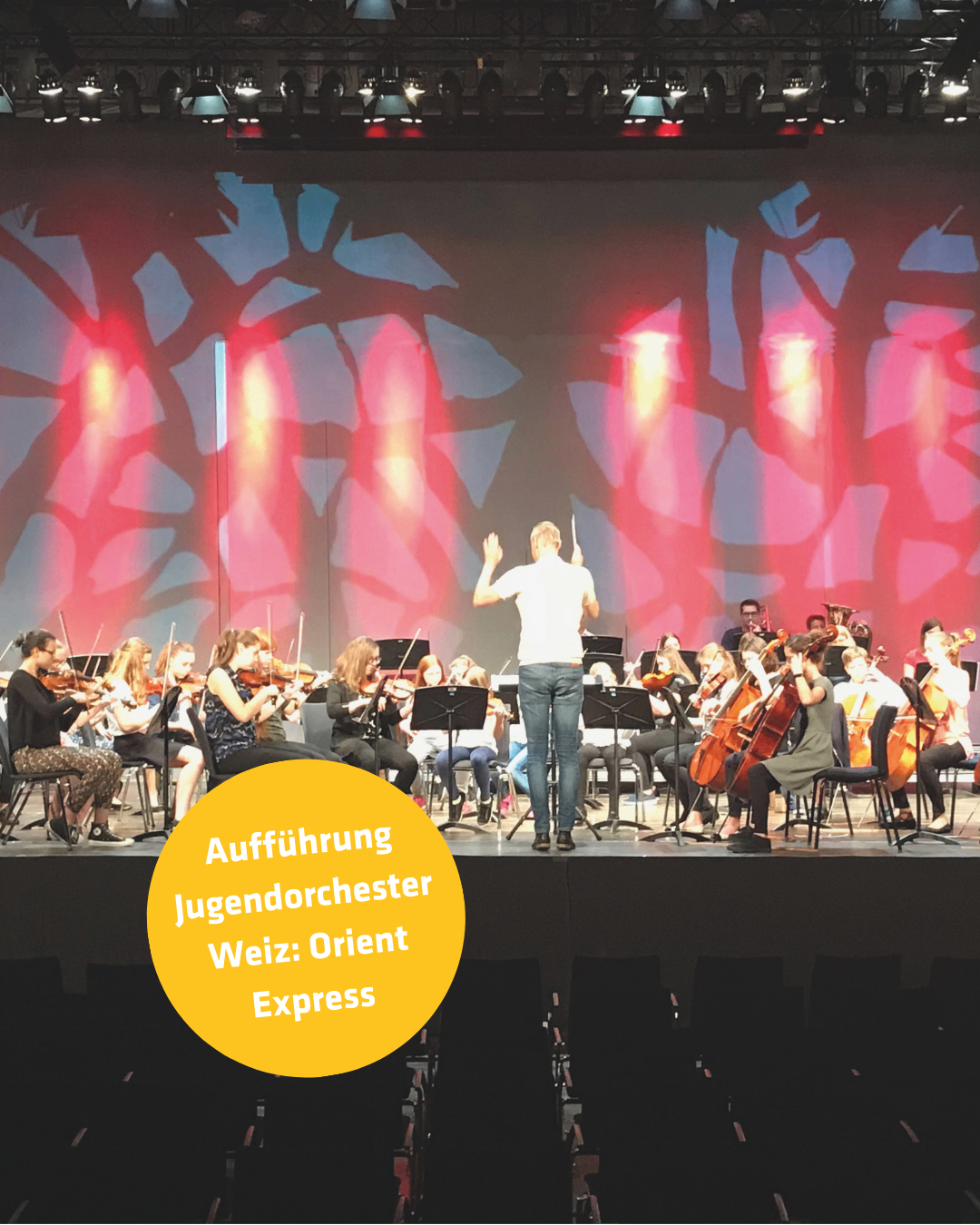 Ein Orchesterauftritt des Jugendorchesters Weiz. Die Bühne ist hell beleuchtet mit roten und orangefarbenen Lichtern. Ein Dirigent leitet das Orchester von der Mitte der Bühne aus.
