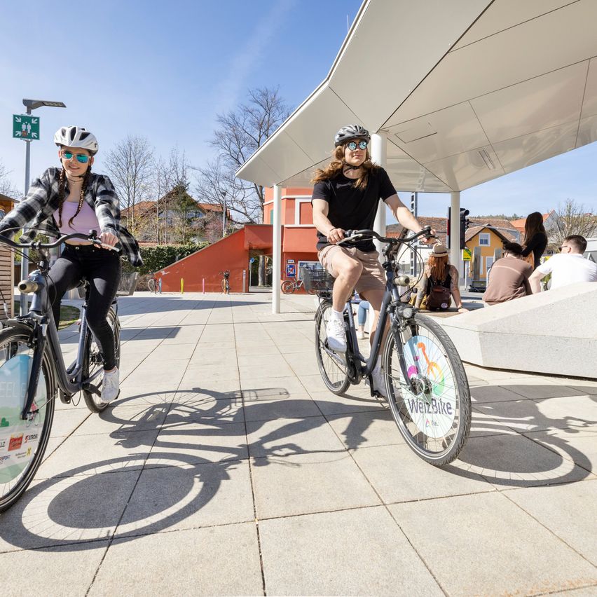 Zwei Radfahrer in Helmen fahren auf einem gepflasterten Bereich in der Nähe einer Fahrradstation. Hinter ihnen sitzen mehrere Personen auf einer Bank. Bäume und Gebäude sind im Hintergrund zu sehen.