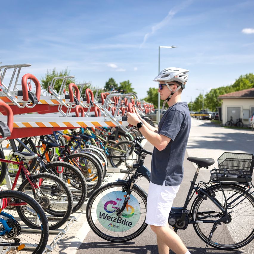 Ein Mann mit Helm und Brille steht neben einer Reihe von Fahrrädern, die in einem Ständer geparkt sind. Die Fahrräder haben ein kreisförmiges Logo mit dem Text 'WeizBike' auf der Vorderseite. Der Himmel ist blau mit einigen Wolken.