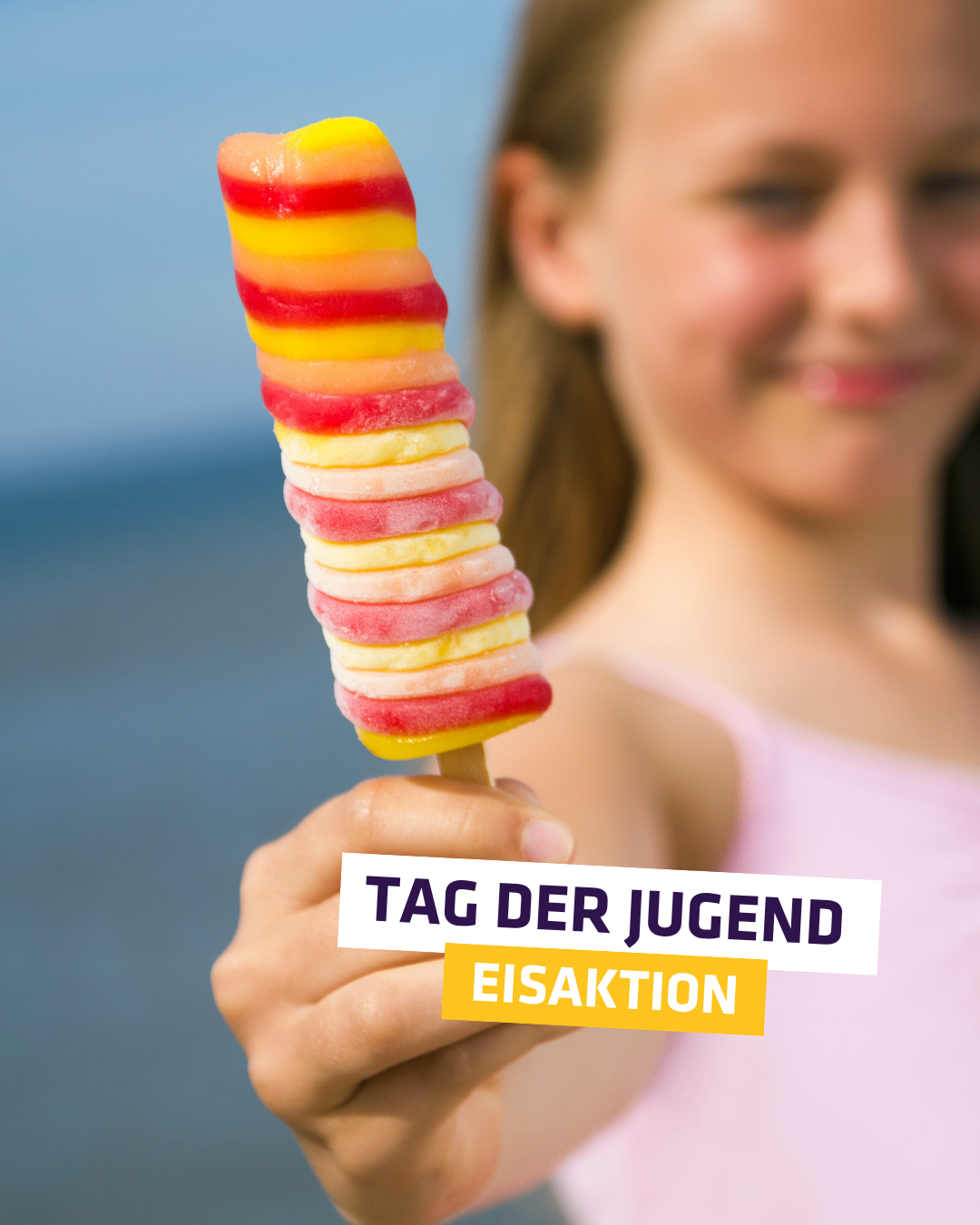 Ein junges Mädchen hält einen mehrfarbigen Eispop am Strand mit einem 'Tag der Jugend' Schild.