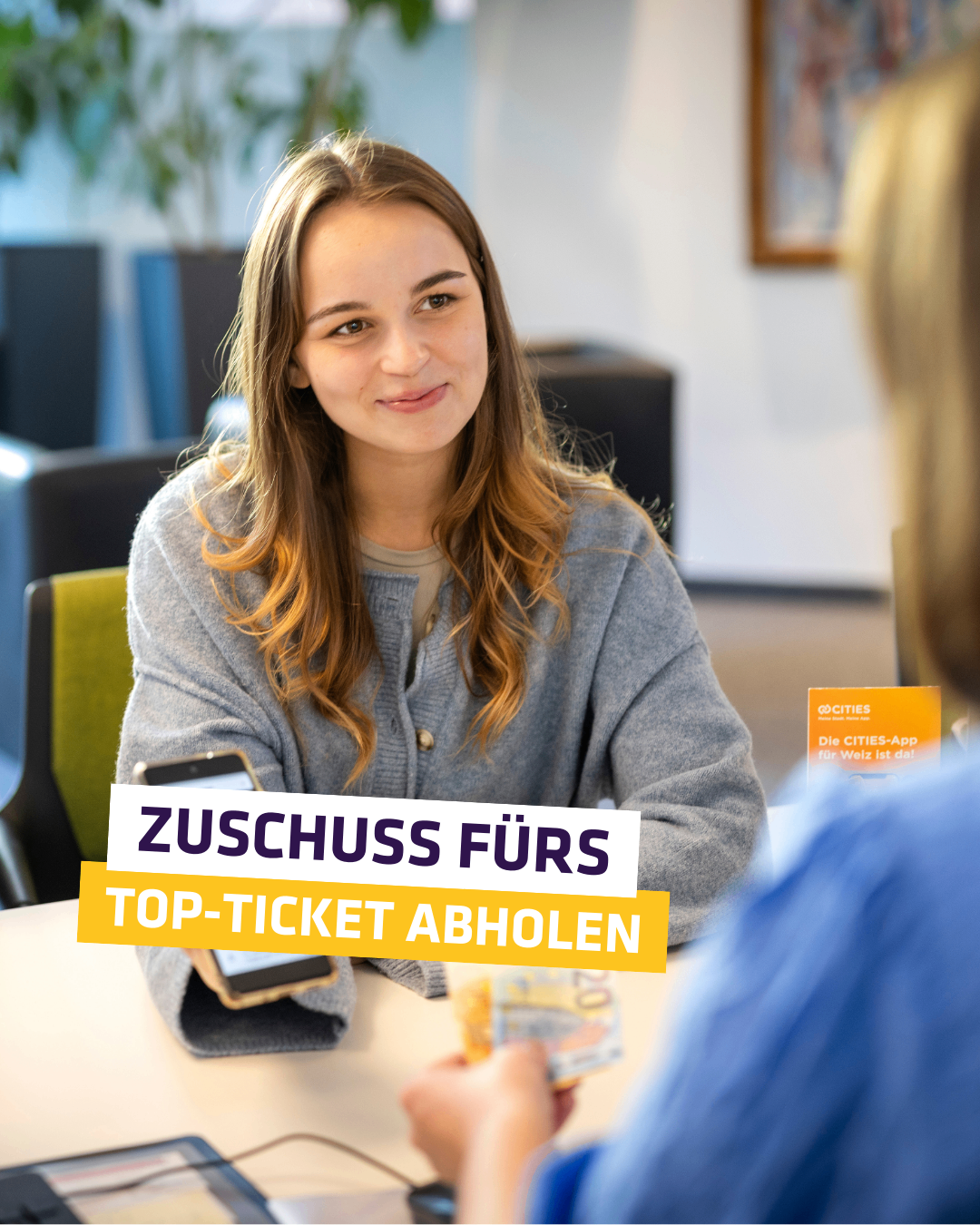 Eine Frau sitzt in einem Büro, lächelt und hält ein Telefon und Geld in der Hand. Sie sieht eine andere Person an, die ebenfalls Geld hält. Ein Schild mit der Aufschrift 'Zuschuss fürs Top-Ticket Abholen' ist vorhanden.