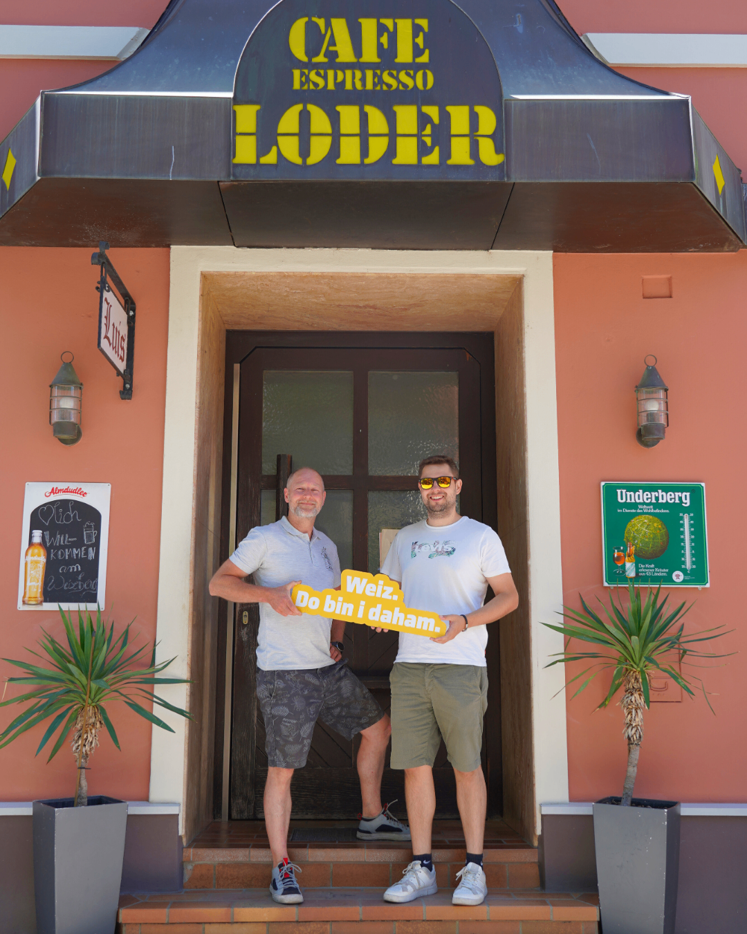 Zwei Männer stehen vor dem Eingang eines Restaurants. Sie halten ein gelbes Schild mit den Worten Weiz. Do bin i daham. Eine Pflanze und eine Lampe sind sichtbar.