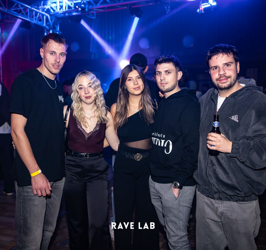 Fünf Personen stehen zusammen in einem Club. Die vier Männer tragen schwarze T-Shirts, eines mit weißer Schrift. Die Frau links hat blonde Haare.