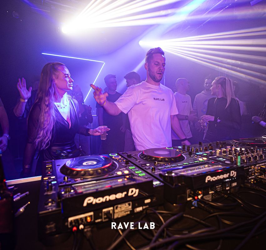 Ein Mann steht hinter Pioneer DJ-Plattenspielern in einem Nachtclub. Eine Frau steht vor ihm und hält ein Getränk.