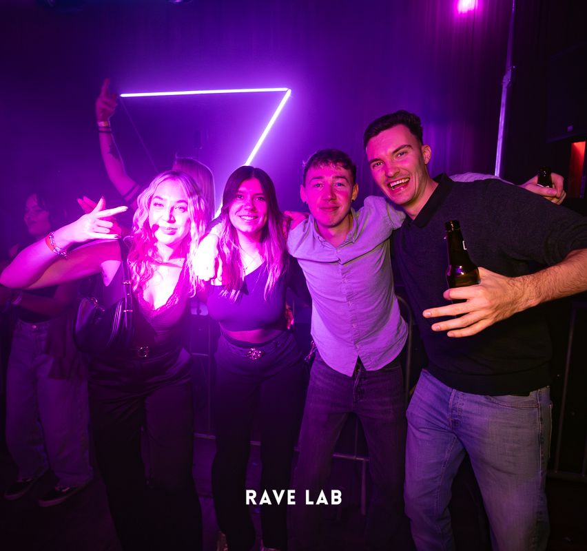 Vier Menschen posieren für ein Foto in einem Club mit lila Licht, zwei Frauen und zwei Männer. Die Männer lächeln, einer hält eine Flasche. Die Frauen haben ihre Hände hochgehoben.