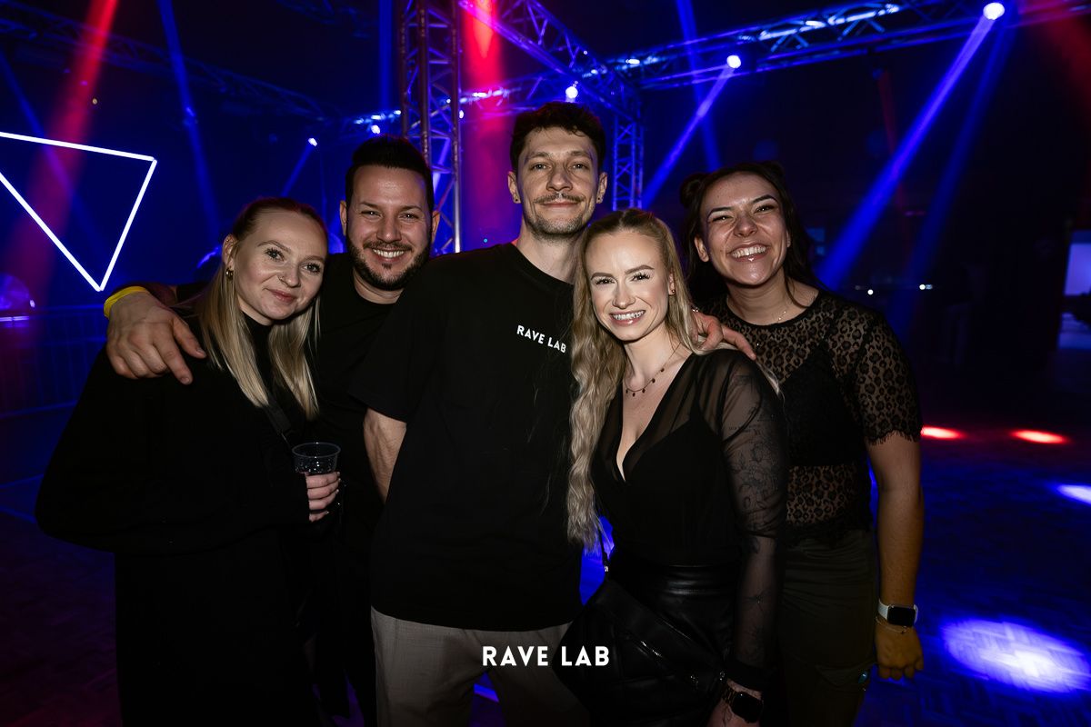 Fünf Personen posieren für ein Foto bei einer Club-Veranstaltung, zwei Frauen und drei Männer. Zwei der Männer tragen Rave Lab T-Shirts. Die Frauen tragen schwarze Kleider. Der Hintergrund hat blaue und rote Beleuchtung.