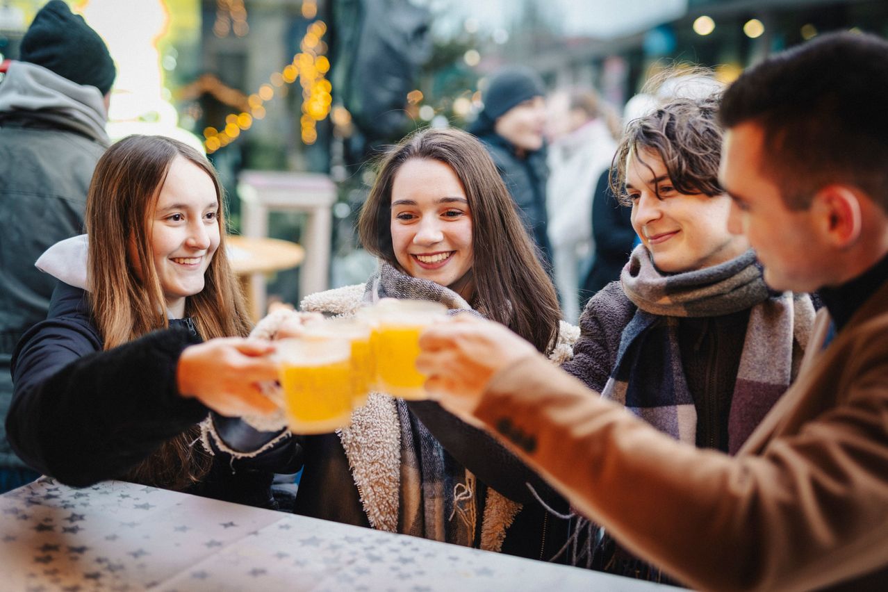 Vier junge Leute, lächelnd, heben Gläser mit gelber Flüssigkeit an einem Tisch an, wahrscheinlich auf einem Festival. Ein unscharfer Hintergrund deutet auf eine festliche Atmosphäre hin.