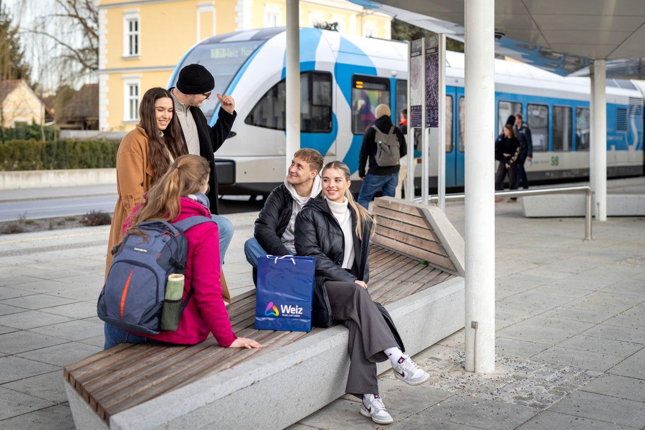 Fünf Personen sitzen auf einer Bank am Bahnhof. Drei lächeln und schauen sich an. Ein blauer Weiz-Taschen liegt zwischen ihnen. Ein Zug ist hinter ihnen geparkt.