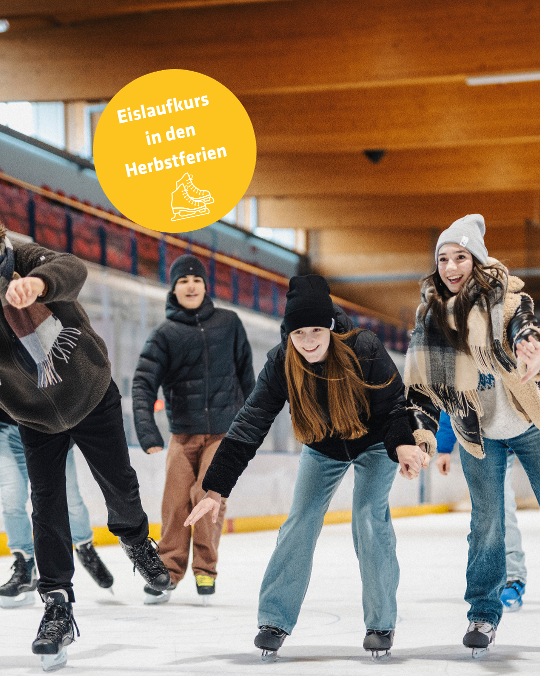 Eine Gruppe junger Leute skatet auf einer überdachten Eisbahn und genießt die Aktivität. Sie tragen Winterkleidung und einige haben Mützen auf. Die Eisbahn hat ein gelbes Schild mit weißem Text, das 'Eislaufkurs in den Herbstferien' sagt.