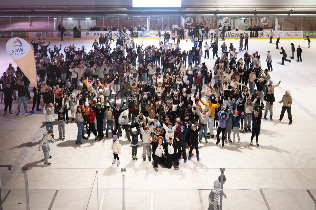 Eine große Menschenmenge, vermutlich eine Schulgruppe, versammelt sich auf einer Eisbahn, in Winterkleidung und teilweise mit Helmen, posiert für ein Foto.