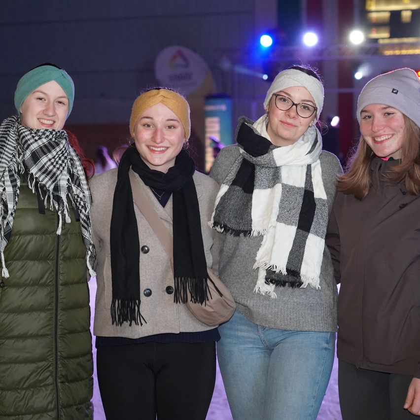 Vier Frauen in Winterkleidung stehen lächelnd zusammen in einem schwach beleuchteten Bereich. Die Frau auf der linken Seite trägt eine grüne Weste. Die Frau neben ihr hat einen Schal mit Karomuster. Die Frau in der Mitte trägt eine Brille.