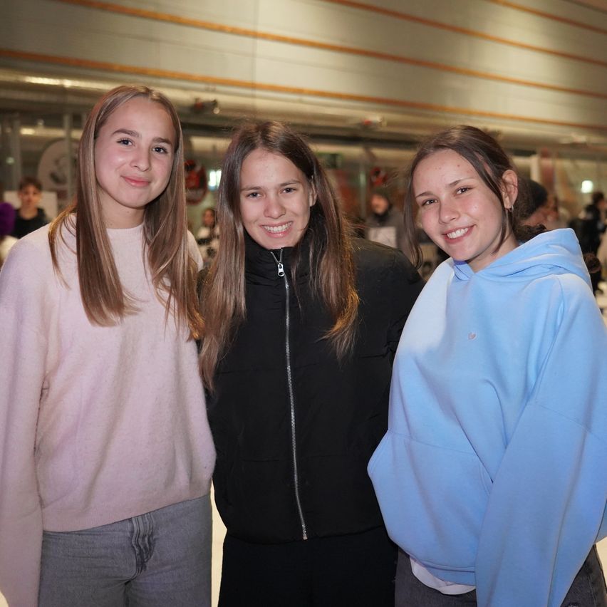 Drei junge Frauen stehen lächelnd zusammen in einem großen Innenraum. Sie tragen verschiedenfarbige Jacken. Im Hintergrund sind Menschen anwesend, einige stehend und andere sitzend.