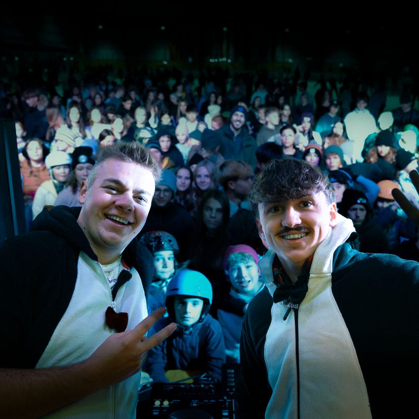 Zwei Männer in Kapuzenjacken posieren für ein Foto vor einer Menschenmenge. Einer hat eine Fliege und der andere einen Schnurrbart. Sie sind von Menschen in Winterkleidung umgeben.