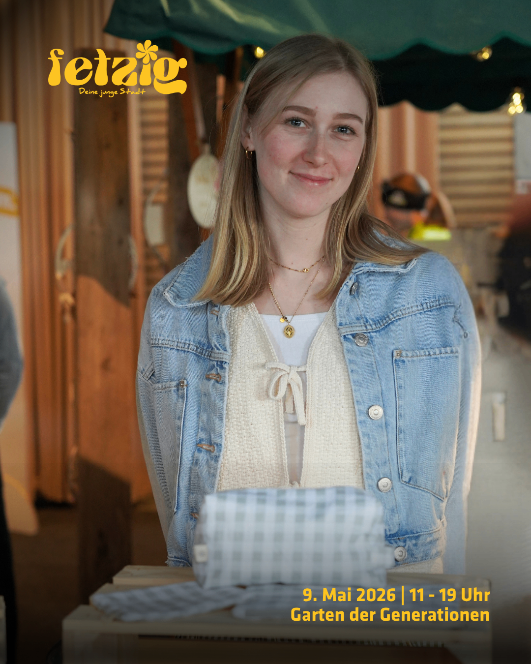 Eine Frau mit blondem Haar, die eine Jeansjacke und eine Halskette trägt, steht vor einer Holzwand mit einer Werbung. Sie hält ein Tuch und lächelt für ein Foto.