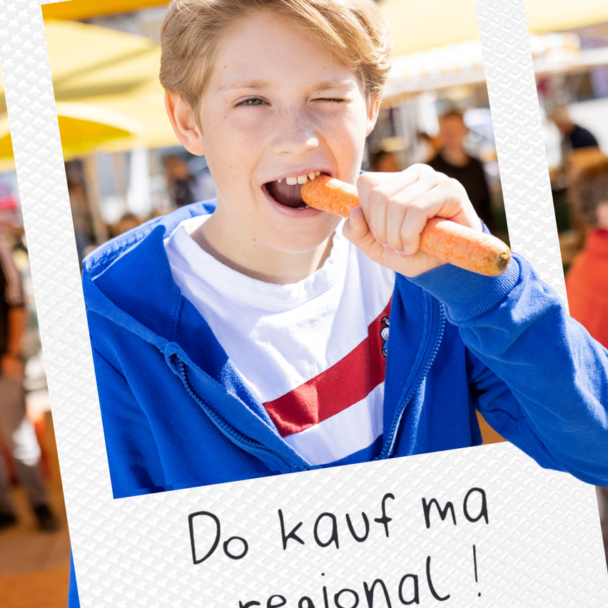 A boy eating a carrot at a market, surrounded by blurry people and umbrellas.