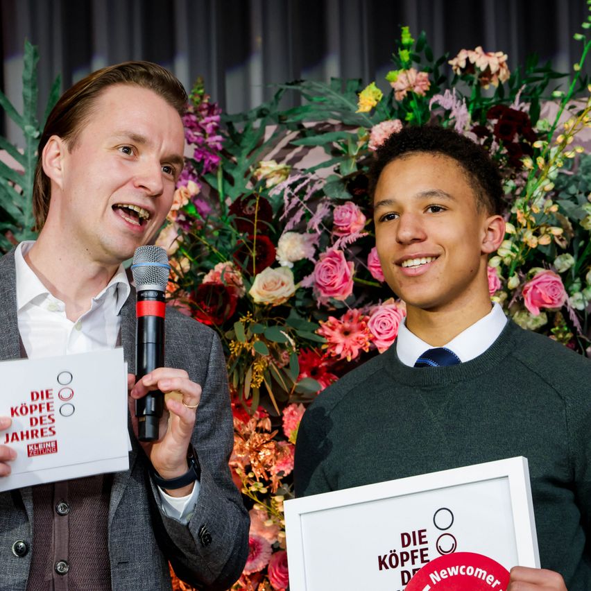 Ein Mann mit Mikrofon und Papier steht neben einem lächelnden jungen Mann, der einen gerahmten Preis hält, umgeben von einem Blumenhintergrund.