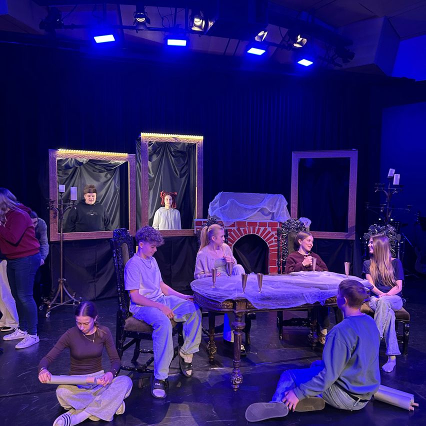 Mehrere Personen sind auf einer Bühne mit blauen Lichtern. Zwei Personen sitzen auf Stühlen, während andere knien. Ein Tisch hat ein Tuch darauf. Es gibt Kerzen und drei große Spiegel an der Wand.