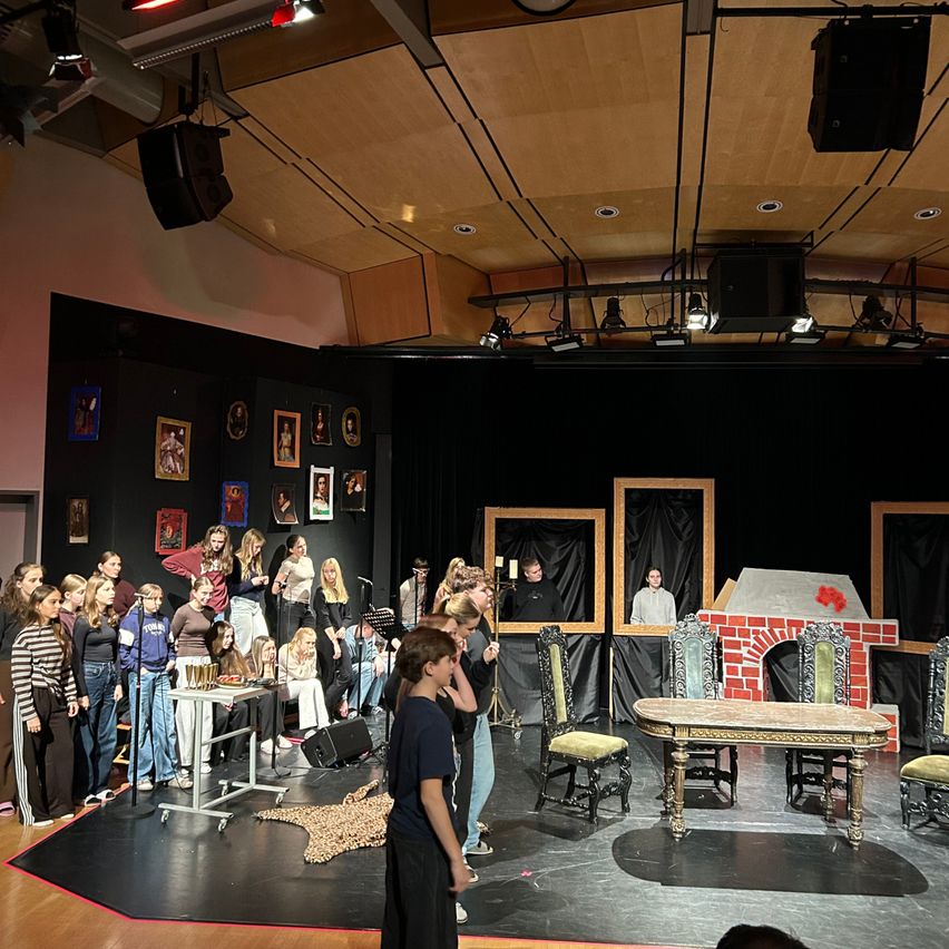 Eine Gruppe von Menschen probt für ein Theaterstück auf einer Bühne. Es gibt Stühle, einen Tisch und einen Kamin. Einige Leute stehen, während andere sitzen. Scheinwerfer und Lautsprecher sind an der Decke.