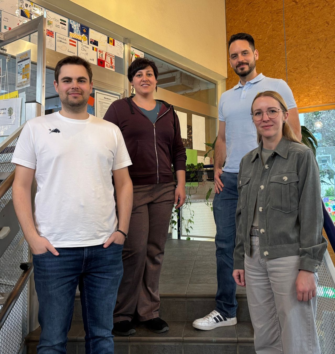 Vier Personen, zwei Männer und zwei Frauen, stehen auf einer Treppe in einem modernen Innenraum. Sie lächeln und posieren für ein Foto. Der Mann auf der linken Seite trägt ein weißes Hemd, und die Frau neben ihm trägt eine braune Jacke.