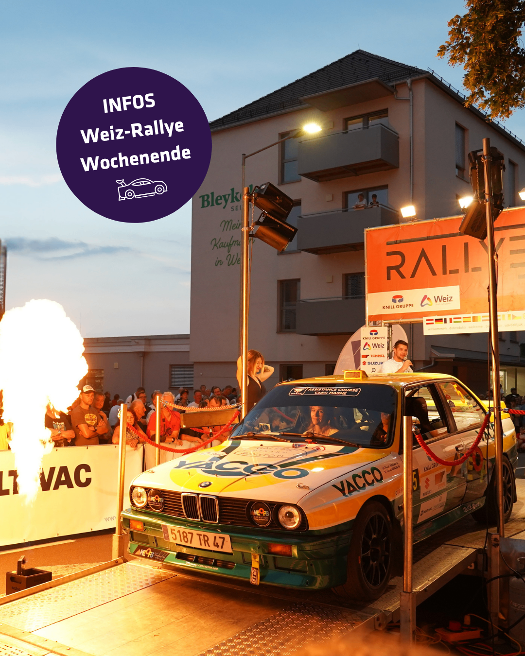 A rally car is on display at an event with spectators and a building in the background. The car has a yellow and green color scheme with the word YACCO on the side. A fire is burning on the left side of the image.