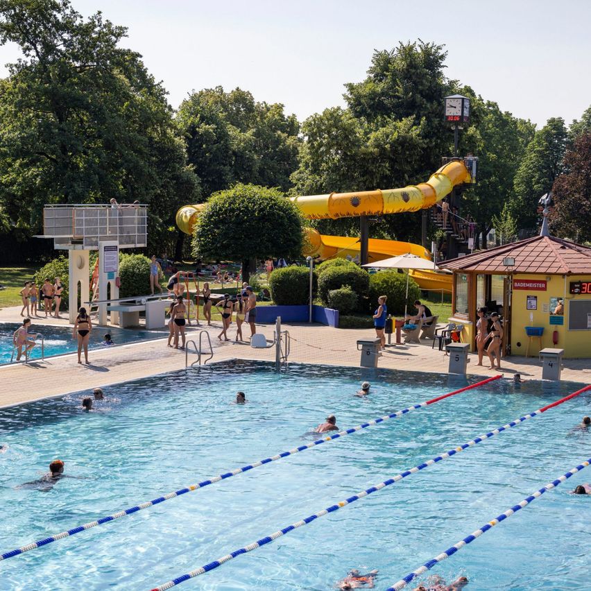 Ein Freibad mit einer gelben Wasserrutsche und Menschen, die in Bahnen schwimmen. Bäume und ein Uhrturm im Hintergrund.