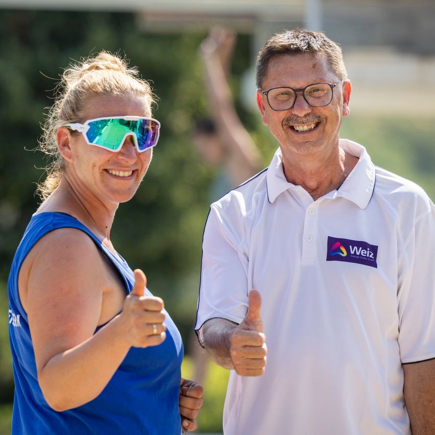 Ein Mann und eine Frau lächeln und geben ein Daumen hoch. Die Frau trägt eine Sonnenbrille und ein blaues Tanktop. Der Mann trägt ein weißes Poloshirt mit Logo. Sie stehen in einem Außenbereich.