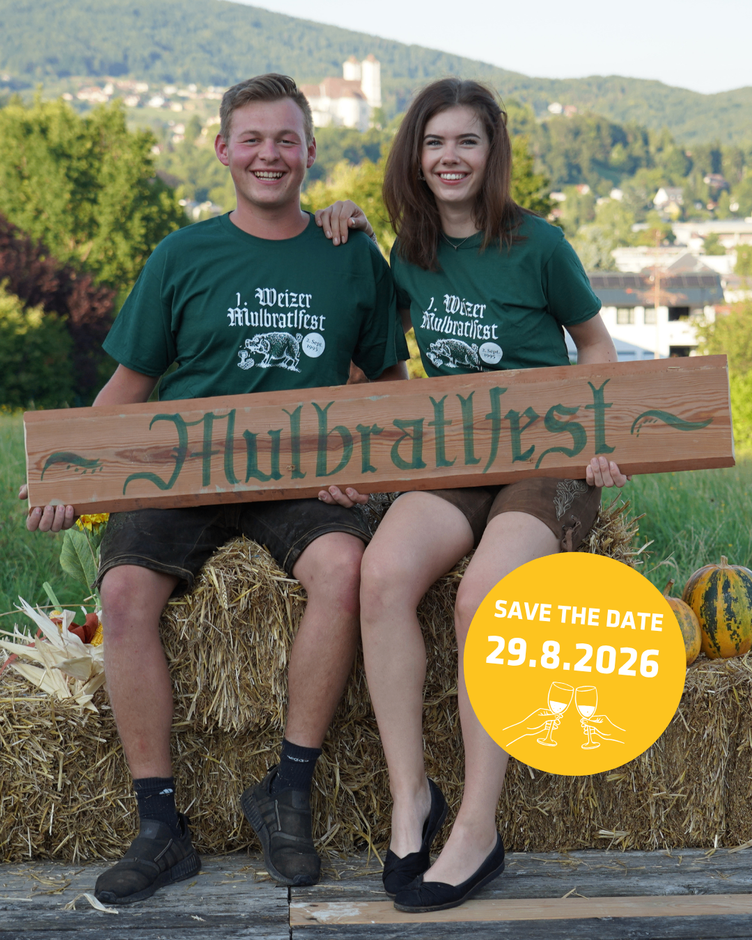Zwei Personen, ein Mann und eine Frau, sitzen auf einem Heuhaufen und halten ein Holzschild mit der Aufschrift 'Hubrattfest'. Sie tragen grüne T-Shirts mit weißem Text und lächeln. Der Hintergrund zeigt eine malerische Aussicht auf Bäume und eine entfernte Stadt.