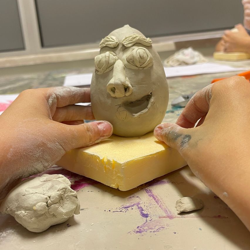 Zwei Hände formen eine Figur aus Ton mit einem Gesicht auf einem gelben Block, bedeckt mit Tonstaub. In der Nähe liegen zusätzliche Tonstücke und Werkzeuge auf einem Tisch.