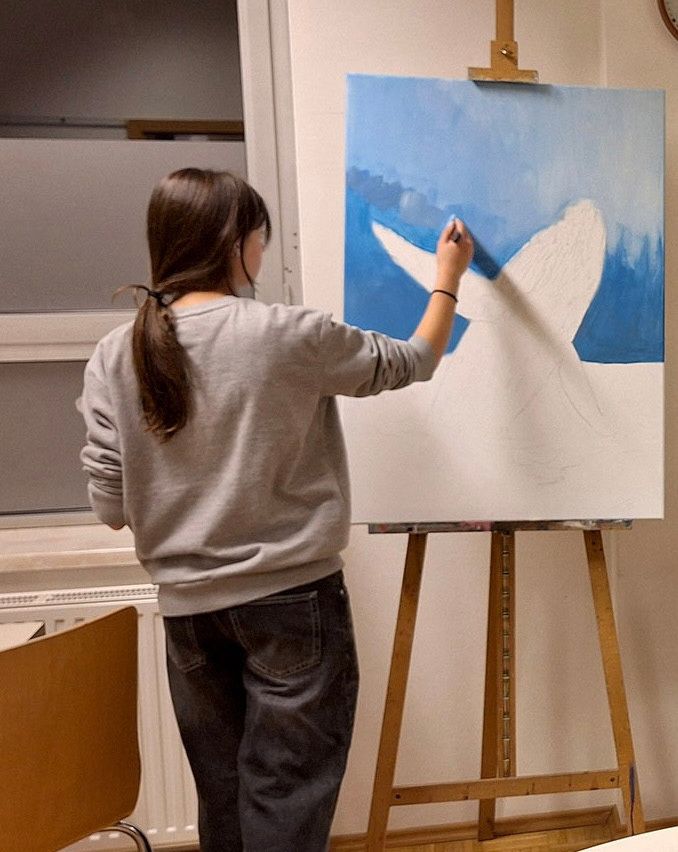 Eine Frau in einem grauen Sweatshirt und Jeans malt einen weißen Vogel auf eine Leinwand mit blauen Farbtönen auf einem Staffelei in einer Innenumgebung.