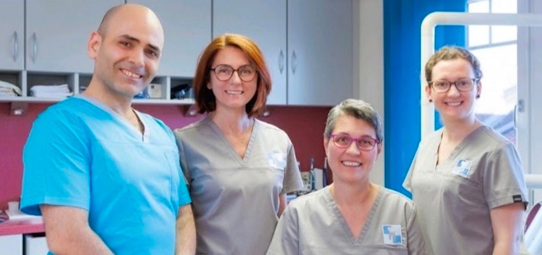 Vier medizinische Mitarbeiter posieren in einer Klinik mit einer blauen Wand hinter ihnen. Sie tragen alle graue Uniformen.