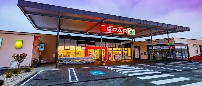 Eingang eines Spar-Geschäfts mit einem violetten Himmel, mit einem roten Schild mit weißen Buchstaben und einem grünen Pfeil. Es gibt einen Parkplatz mit einem Zebrastreifen.