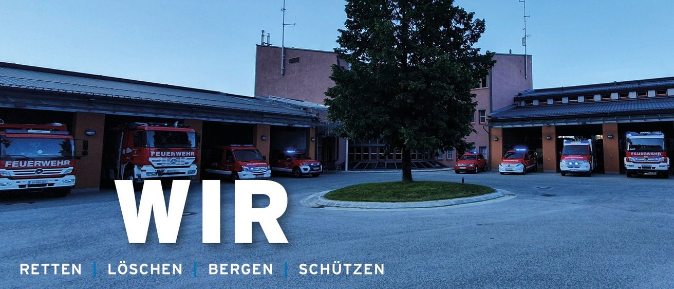 Ein Feuerwehrhaus mit roten Rettungsfahrzeugen davor. Ein Baum steht in der Mitte, mit Gebäuden auf beiden Seiten. Der Himmel ist klar und blau.