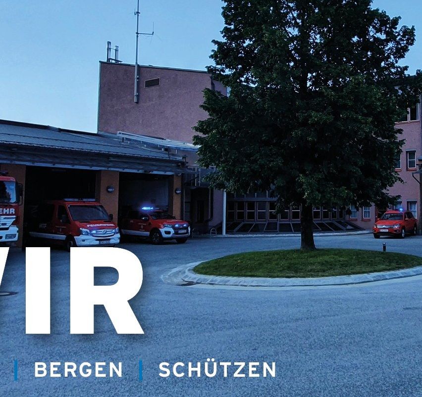Ein Feuerwehrhaus mit roten Rettungsfahrzeugen davor. Ein Baum steht in der Mitte, mit Gebäuden auf beiden Seiten. Der Himmel ist klar und blau.