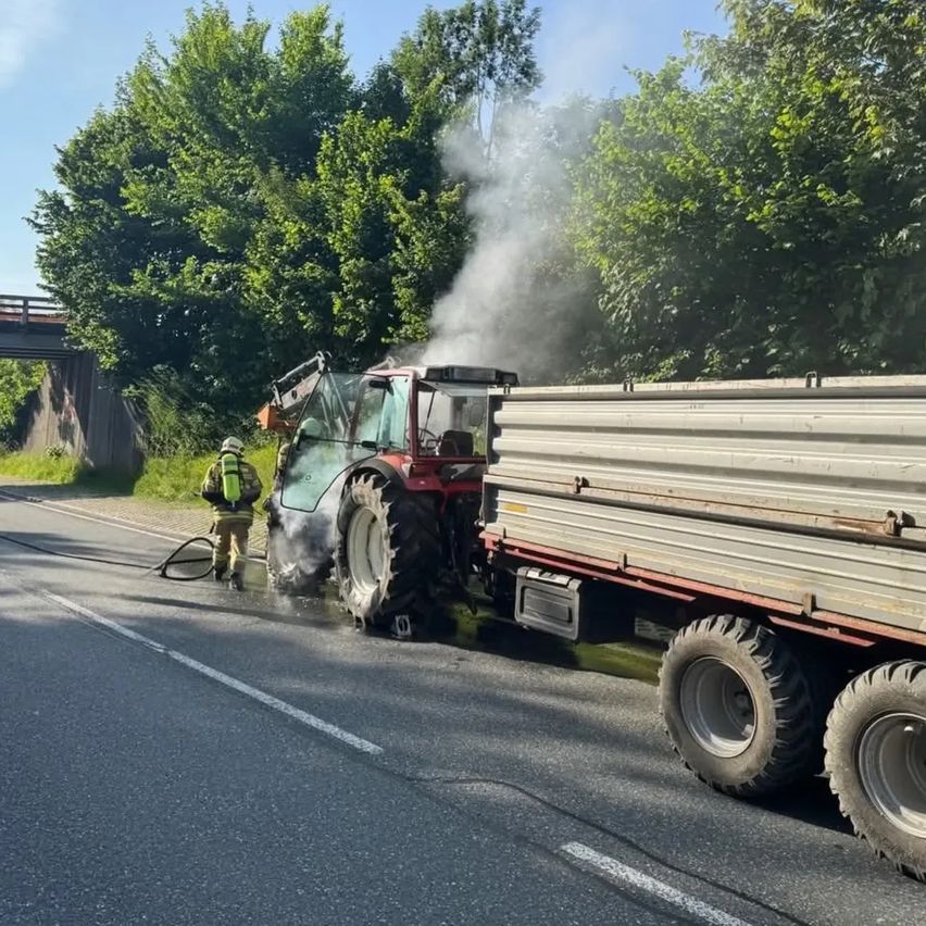 Ein Feuerwehrmann sprüht mit einem Schlauch Wasser auf einen rauchenden Traktor und Anhänger am Straßenrand, mit Bäumen und einer Brücke im Hintergrund.