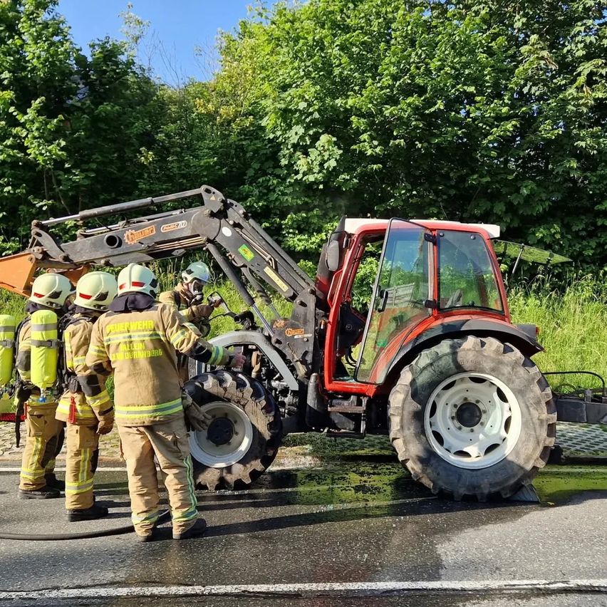 Feuerwehrleute in voller Ausrüstung kümmern sich um einen roten Traktor am Straßenrand. Im Hintergrund befinden sich Bäume und Büsche.