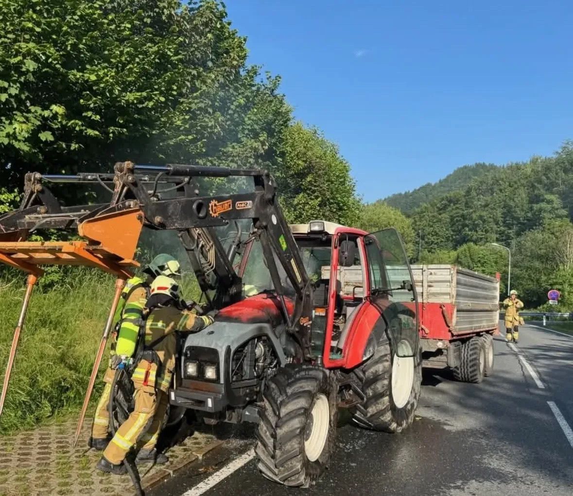 Zwei Feuerwehrleute arbeiten an einem roten Traktor und Anhänger am Straßenrand, mit einer Person, die am Straßenrand geht, und einer Straßenlaterne in der Nähe.