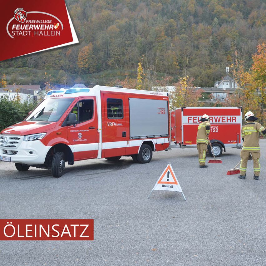 Ein Feuerwehrauto ist geparkt, während zwei Feuerwehrleute den Bereich reinigen. Ein Warnschild mit Ausrufezeichen steht auf der Straße.