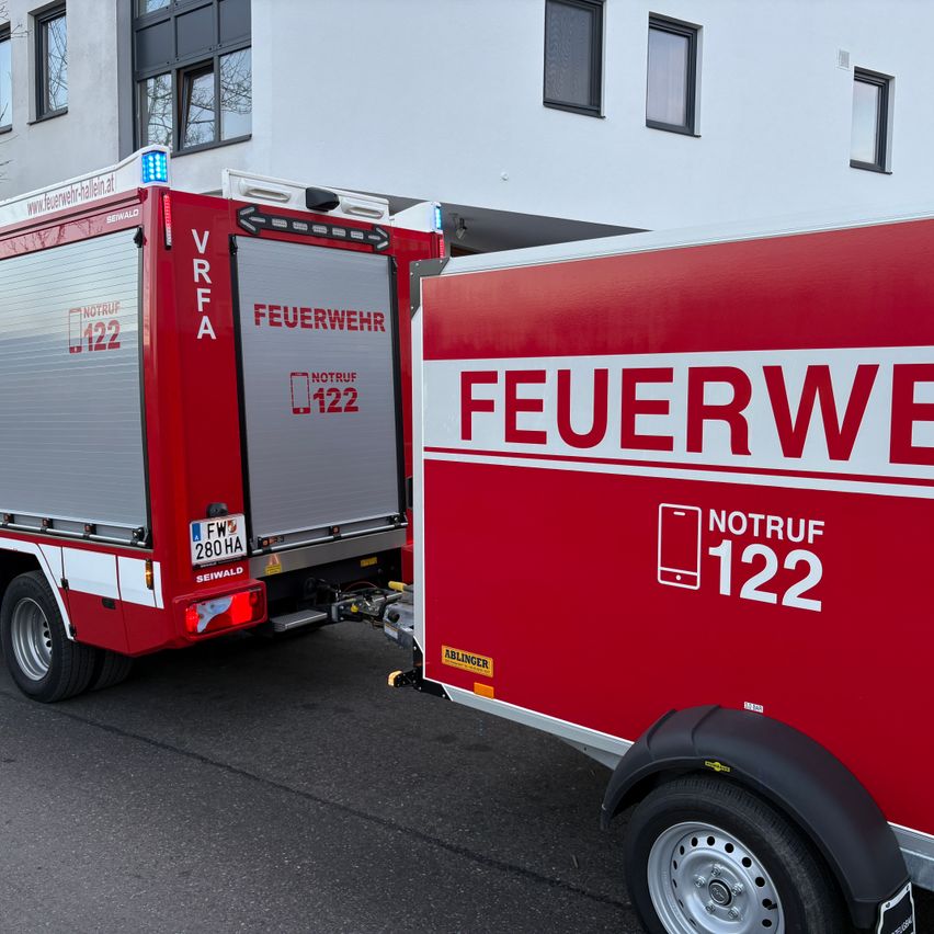 Zwei rote Feuerwehrfahrzeuge sind vor einem weissen Gebäude mit mehreren Fenstern geparkt. Die Wörter NOTRUF und die Nummer 122 sind auf den Fahrzeugen geschrieben.